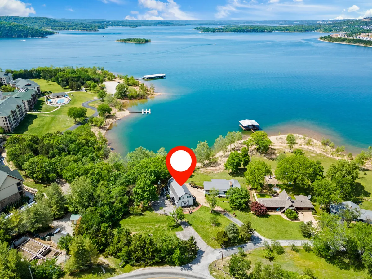 A wide-angle view of the lakefront, featuring a shared dock extending into the water, the house, and lush surrounding greenery. The scene includes space for swimming, boating, fishing, and relaxing by the water, with a backdrop of tranquil sunset lighting.