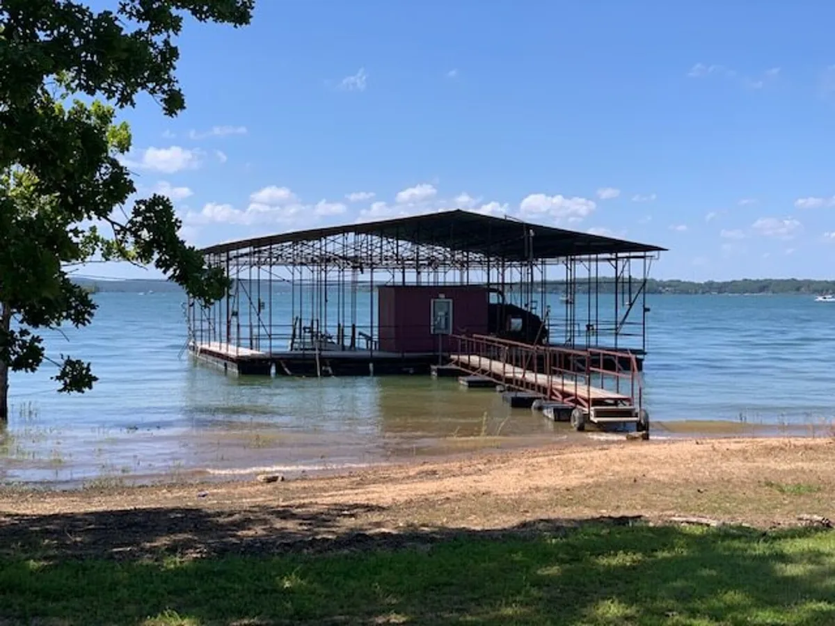 A serene lakeside scene featuring a wooden dock extending into calm waters, flanked by lush trees. A small boat is tied to the dock, and the shoreline invites families to fish, swim, or launch kayaks. The setting sun casts a warm glow over the water, enhancing the welcoming and tranquil mood.