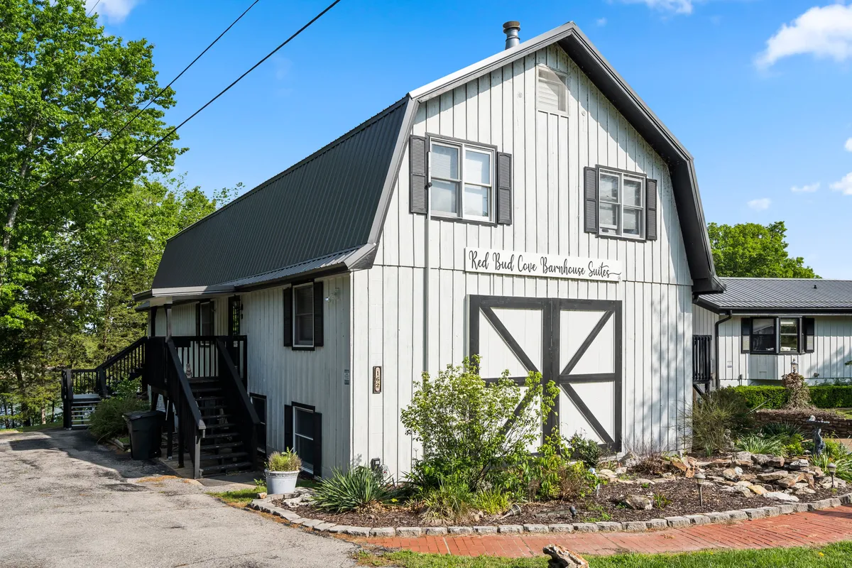 An exterior view of the barn-style suite, featuring a landscaped garden and welcoming porch with visible signage. The inviting scene suggests opportunities for enjoying morning coffee outdoors or gathering in the evening.