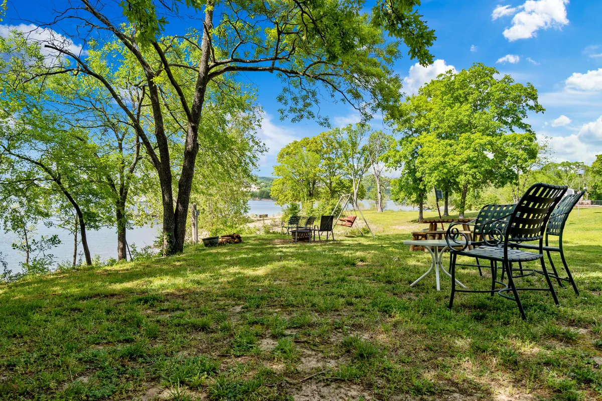 A peaceful lakeside lawn with relaxed lawn chairs facing the water, surrounded by trees and a swing set. The scene is set for quiet moments, conversations, and enjoying the natural setting, with the lake stretching into the distance.