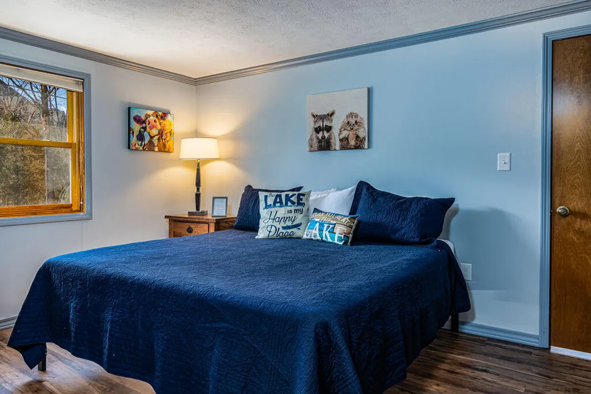 This image showcases a serene bedroom with a neatly made bed dressed in decorative pillows, a bedside lamp, and a window that lets in soft natural light. The décor is warm and inviting, hinting at a cozy retreat ideal for rest and relaxation.