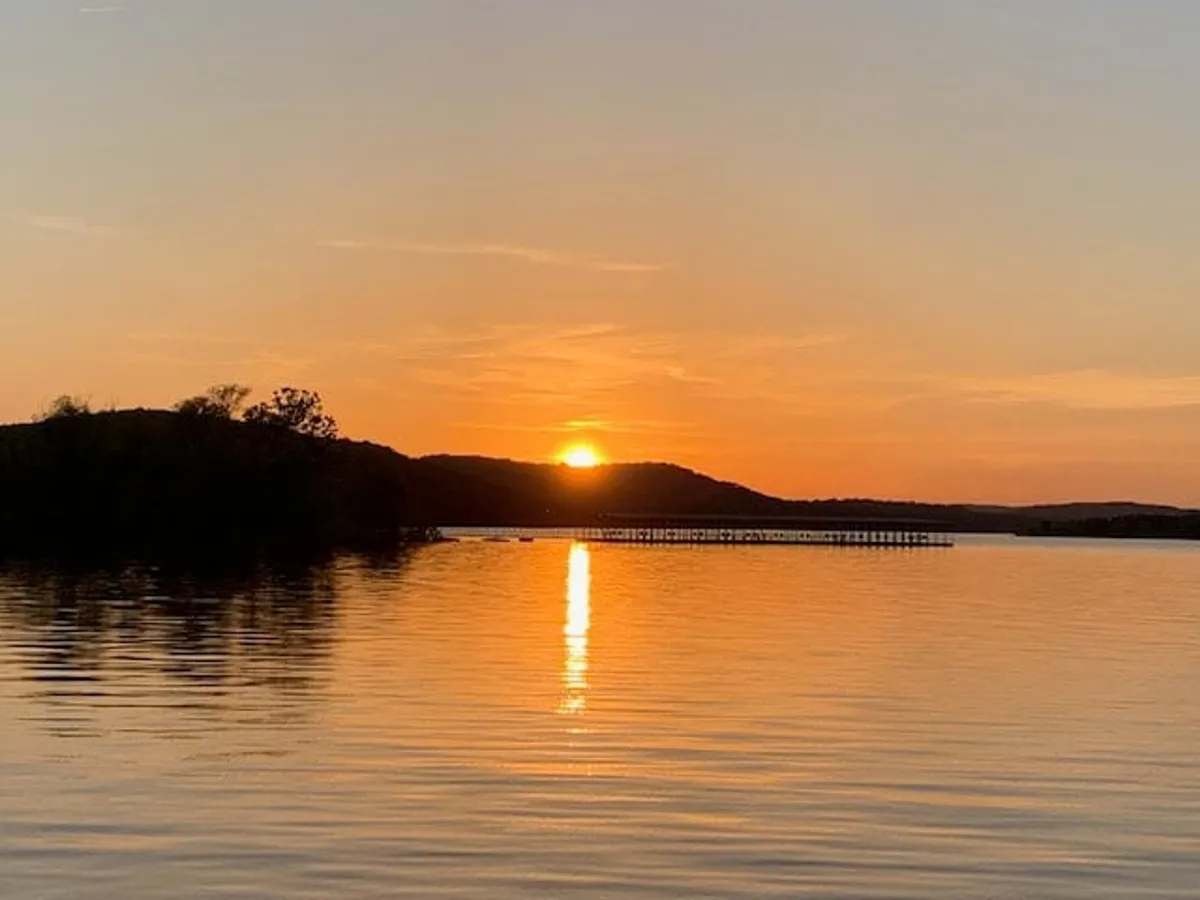 A tranquil scene of the sun setting on the horizon, with its warm colors reflecting off the calm water. Silhouetted hills and a pier extending into the lake evoke a sense of peace and connection with nature, ideal for evening relaxation.