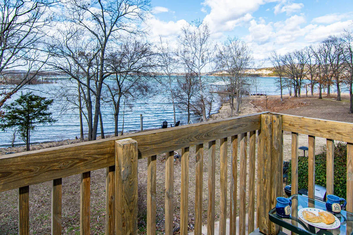 An exterior shot from the deck looking out over the lake, with a table set for outdoor dining, surrounded by trees and featuring a tranquil water view. The deck railing frames the scene, capturing both the comfort of the outdoor seating and the natural setting.