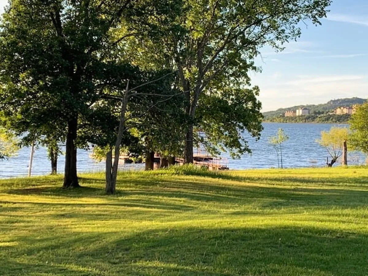 A peaceful exterior view of large trees shading a grassy area that gently slopes toward the lake. Distant buildings and boats are visible on the water, suggesting a communal, family-friendly space for picnicking, fishing, and boating. The natural setting is ideal for gatherings and enjoying nature.