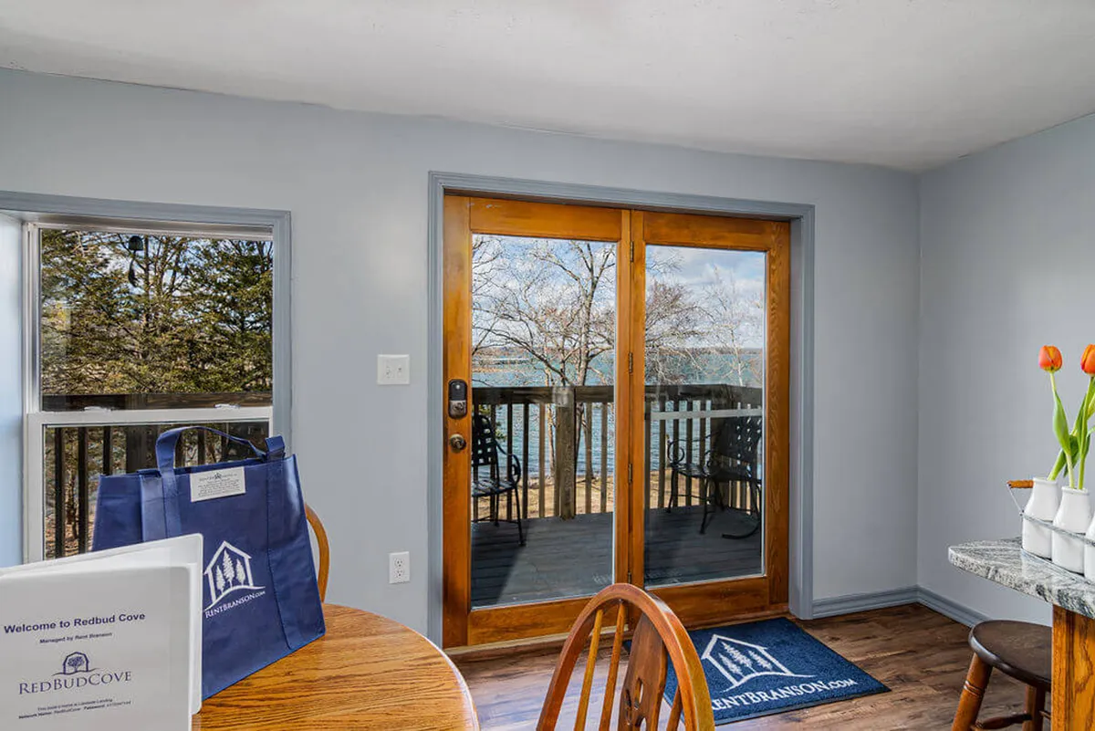 An interior view with sliding glass doors opening to a wooden deck that overlooks the lake. A table with tulips sits near the doors, highlighting a perfect spot to enjoy meals or coffee while soaking in the scenic beauty. The scene is serene and welcoming, blending indoor and outdoor relaxation.