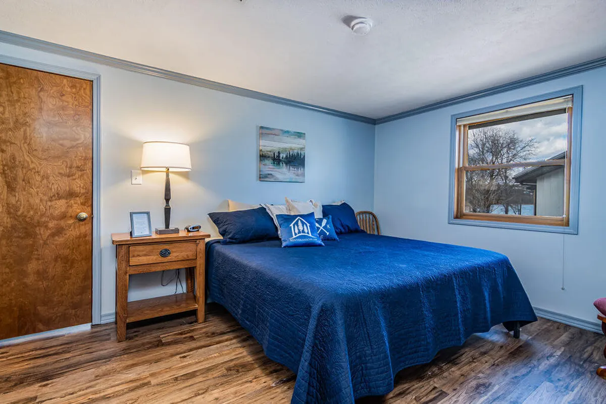A relaxing bedroom scene featuring a bed with blue bedding, a wooden nightstand with a lamp, framed artwork, and a window offering a glimpse of the outdoors. The room feels peaceful and updated, perfect for a restful stay.