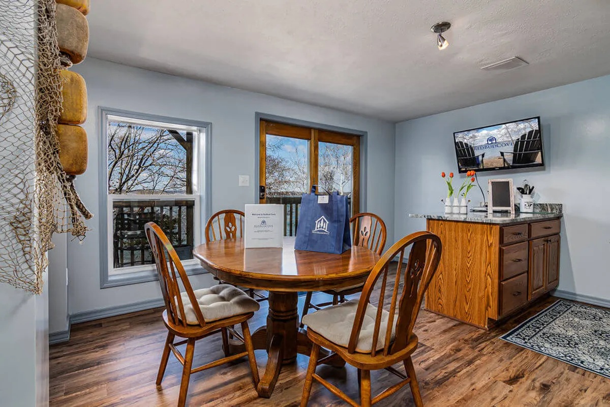 The image showcases a sunlit living and dining area with a round wooden table surrounded by four chairs, large windows overlooking Table Rock Lake, a kitchenette, and a television. The space is bright and welcoming, inviting guests to gather, dine, and enjoy the lake views from inside.